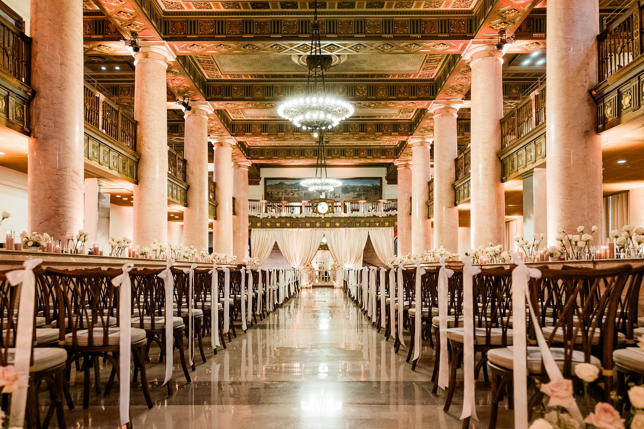 Elegant wedding reception hall with tall pink marble columns, grand chandeliers, and long dining tables decorated with white flowers and ribbons.