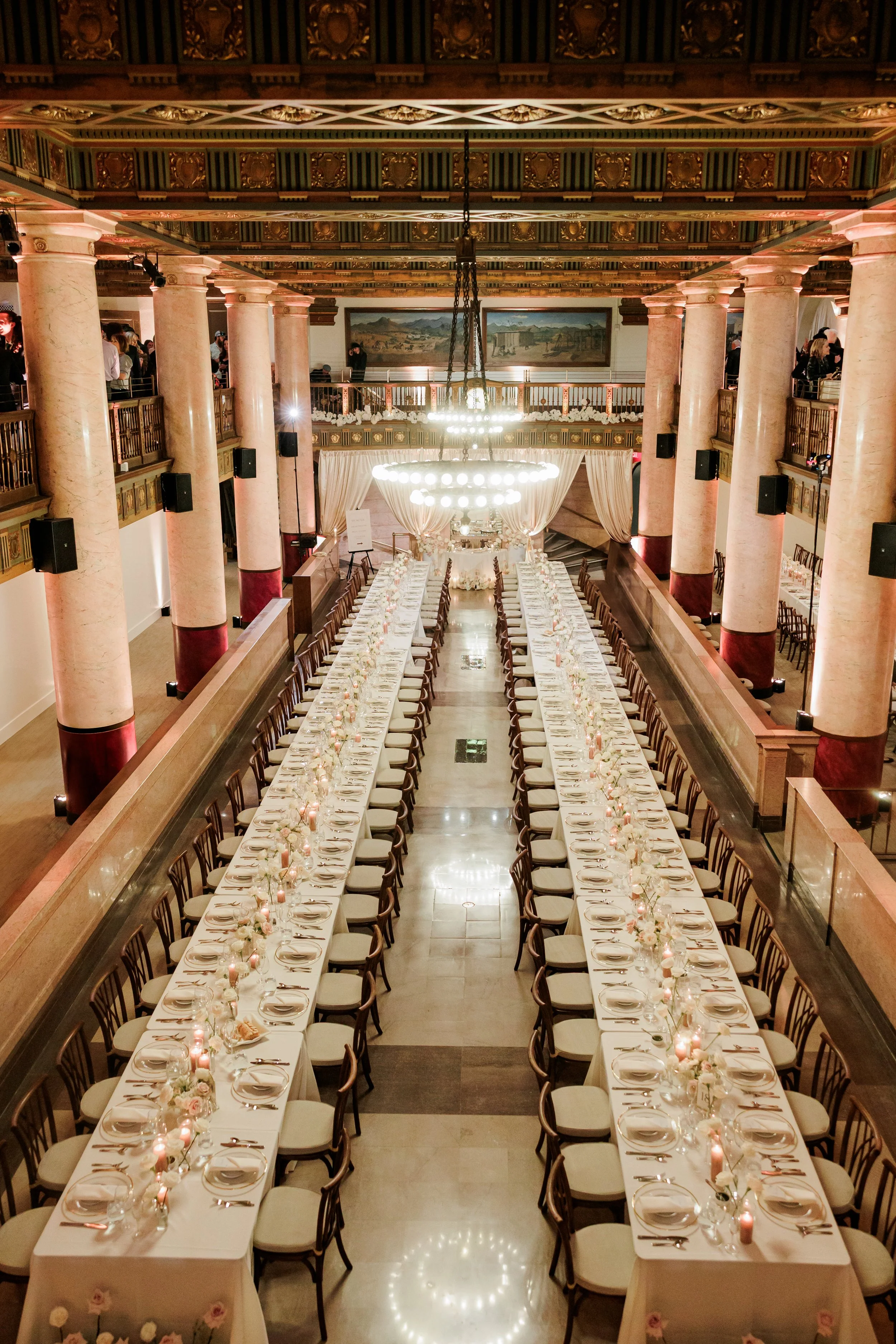 Elegant banquet hall with long tables set for a formal event, decorated with flowers and candles, surrounded by chairs, with large columns, a chandelier, and a balcony.