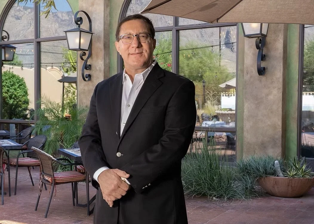 A man in a black suit and white shirt standing outdoors at a restaurant or cafe with patio furniture and large windows.