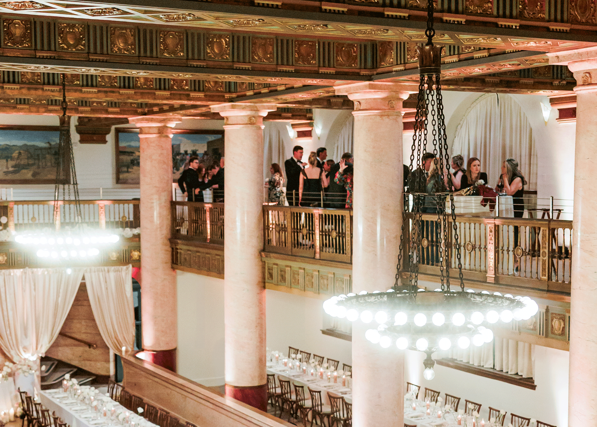 An elegant banquet hall with tall pink marble columns, ornate ceiling details, and a balcony area with people socializing. The lower level has a long dining table set for a formal event, with candles and floral centerpieces, all illuminated by a grand chandelier.