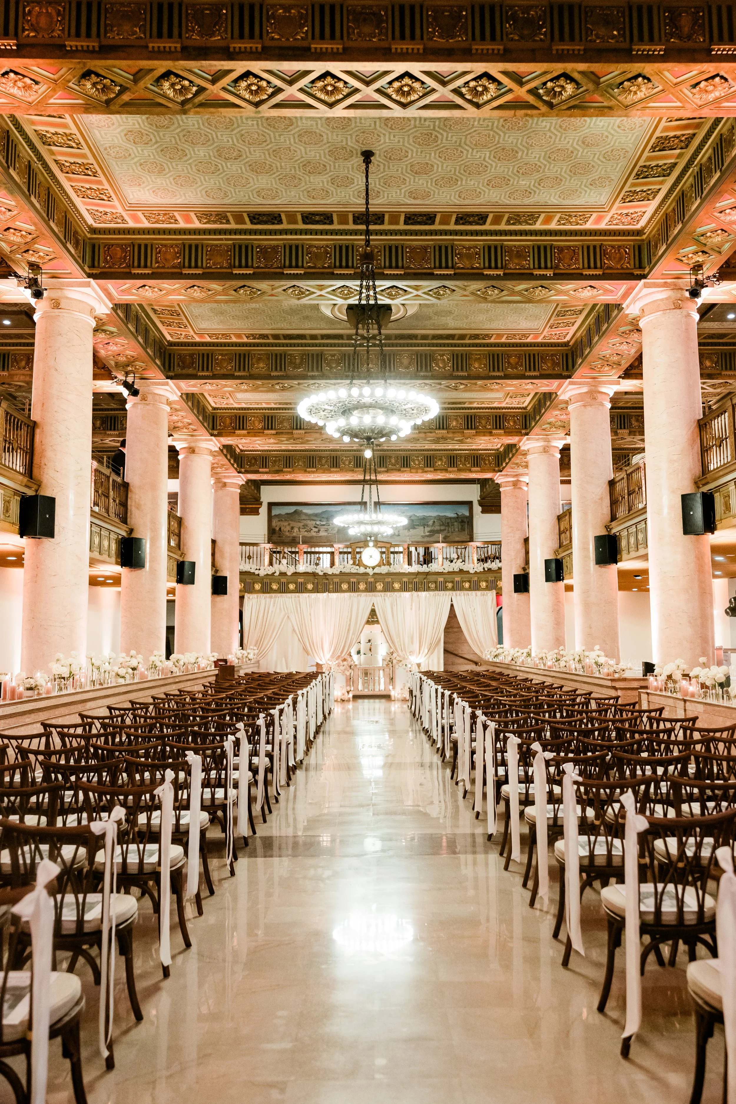 Elegant indoor venue set up for a wedding ceremony with rows of chairs, floral arrangements, and draped curtains at the altar, in a hall with pink columns, ornate ceiling, and chandeliers.