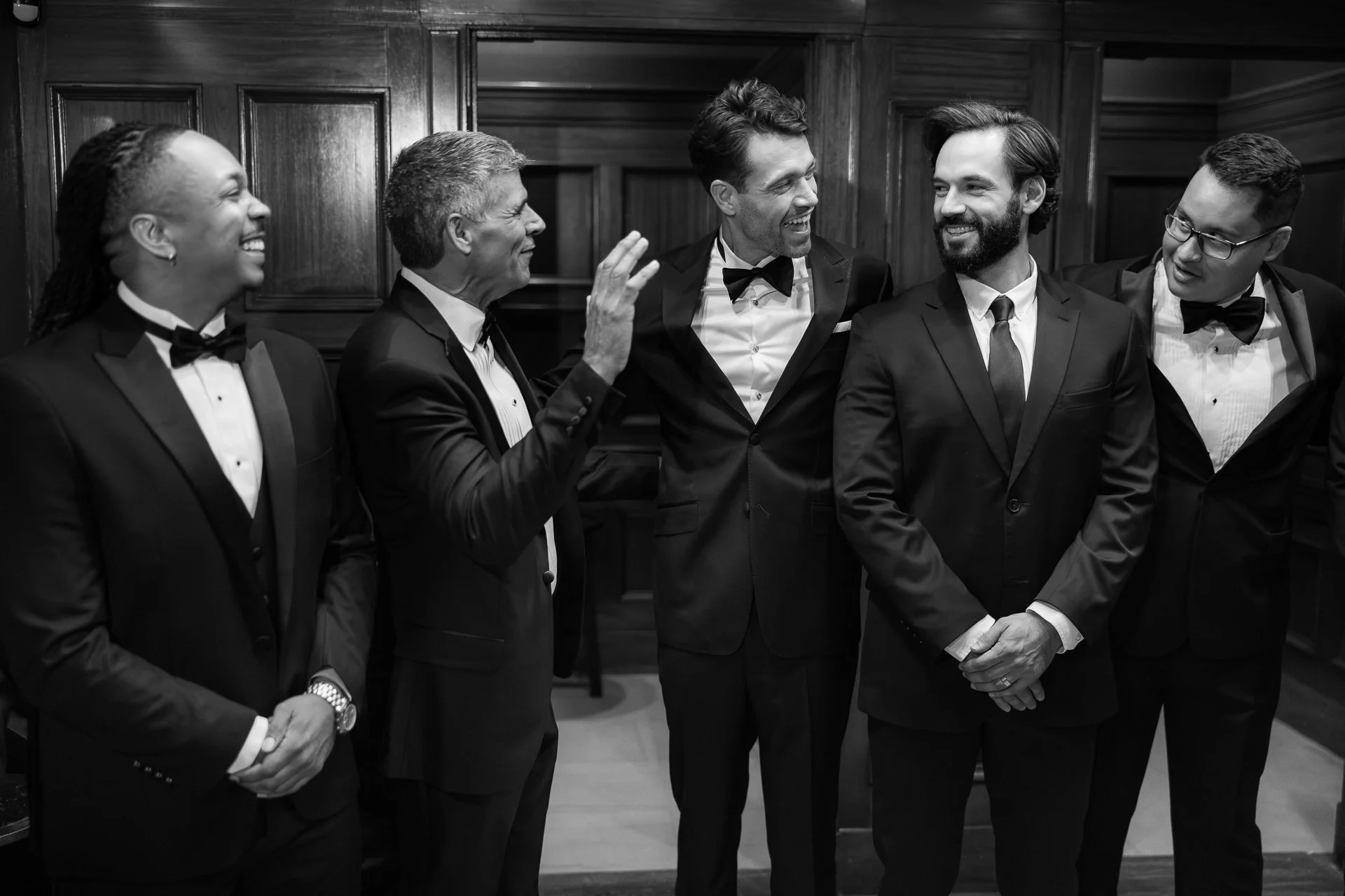 Six well-dressed people in tuxedos engaged in conversation, smiling, standing in a room with wooden paneling.