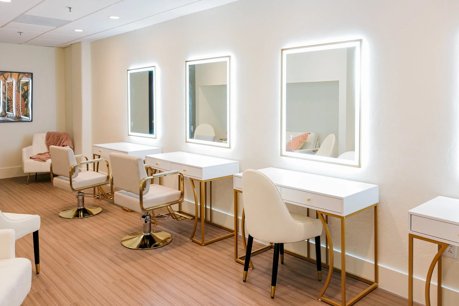 Beauty salon station with white chairs, illuminated mirrors, and white tables with gold accents.