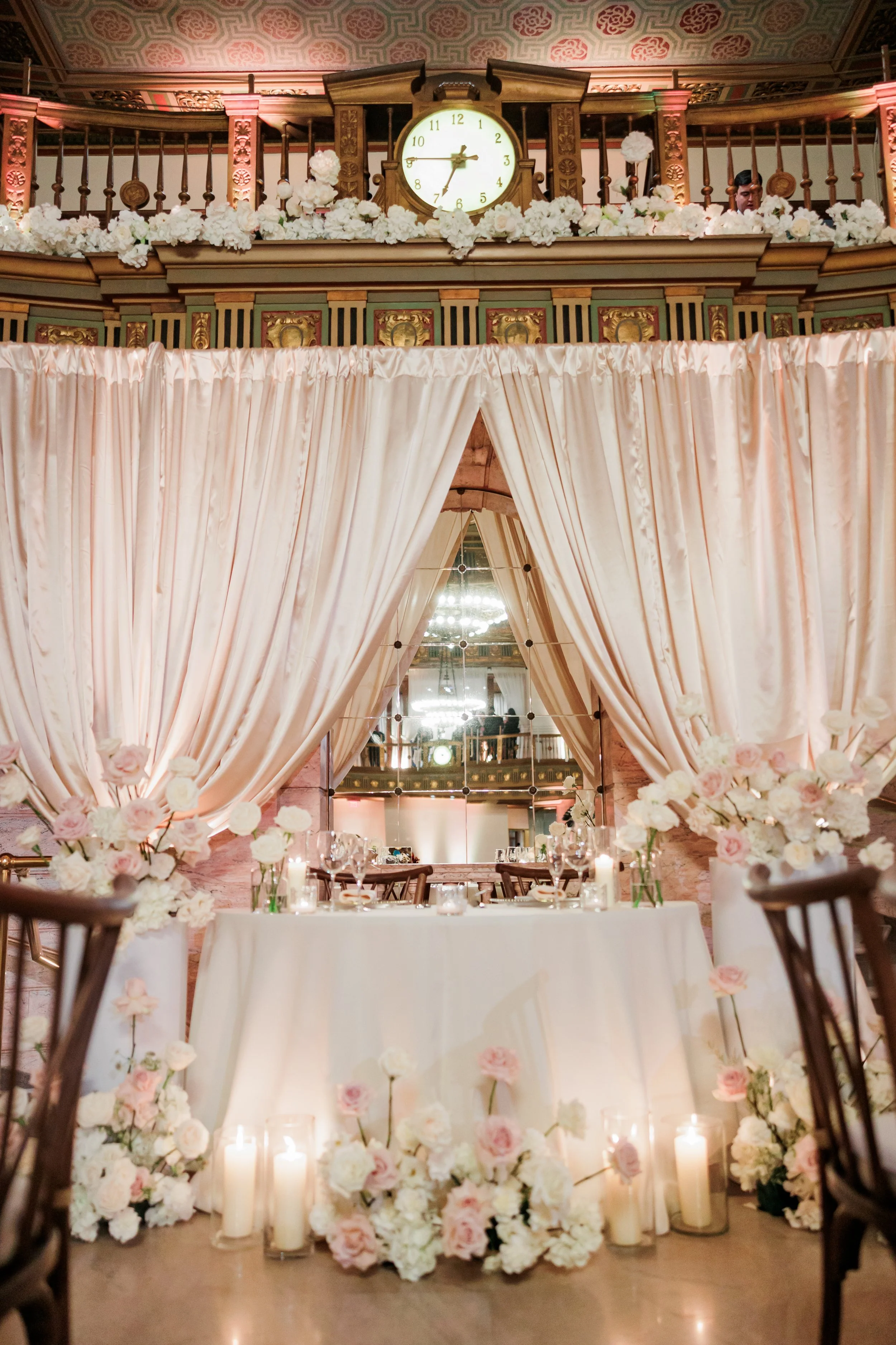 Elegant wedding reception setup with a decorated head table, pink and white flowers, candles, and draped curtains in a historic venue with a clock and ornate woodwork.