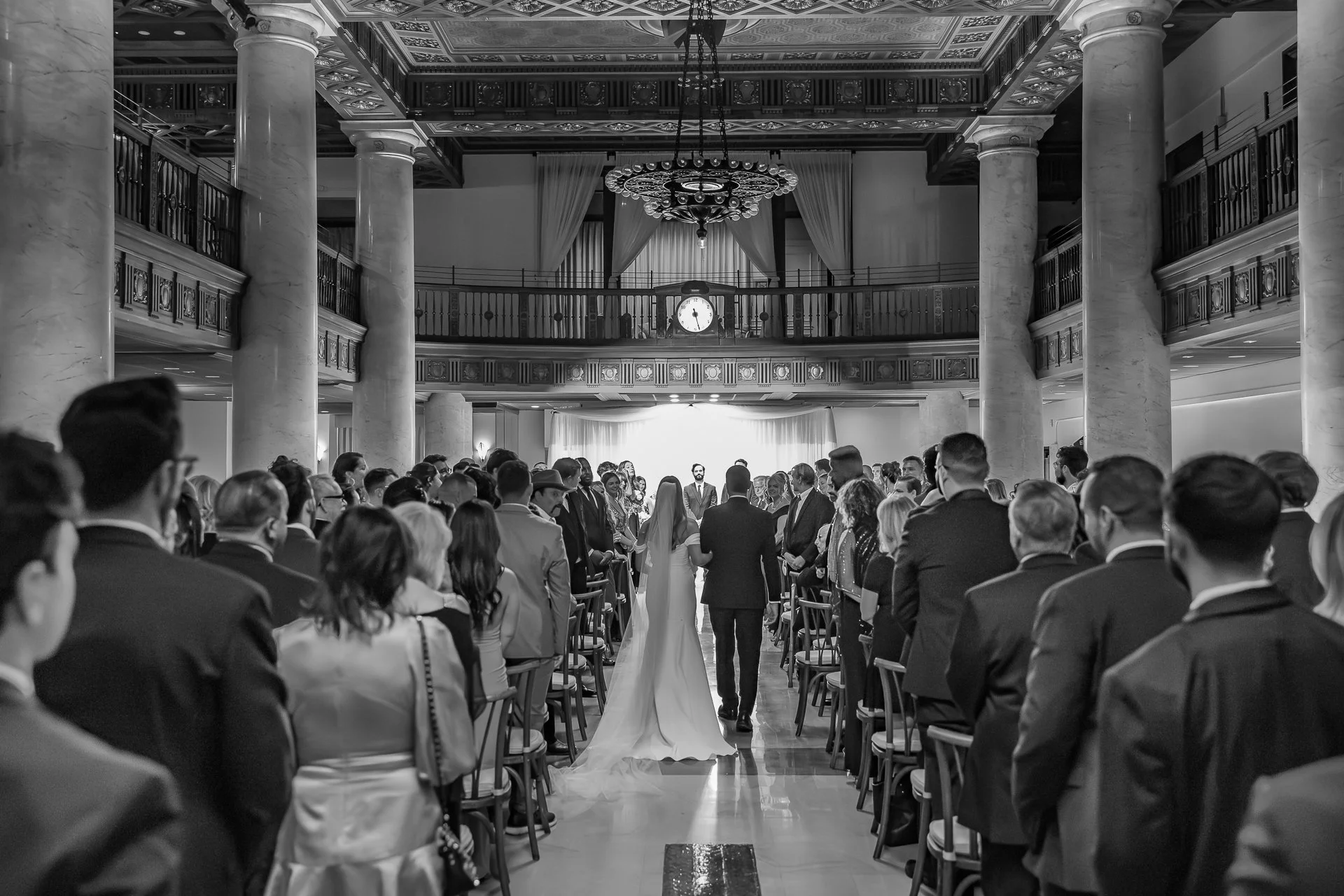 Black and white photo of a wedding ceremony inside a grand hall with many guests, two people walking down the aisle towards the altar.