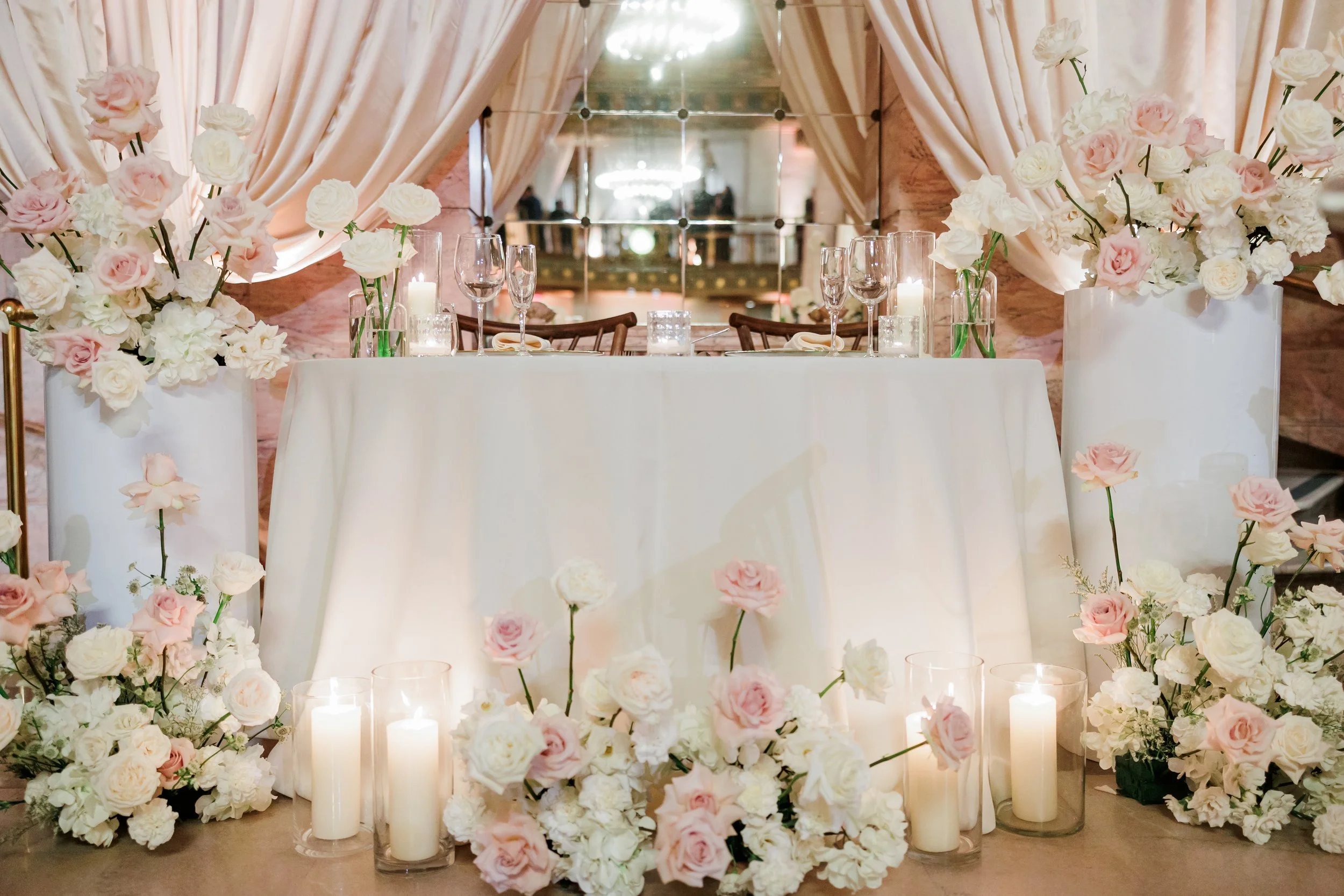 Elegant wedding reception table decorated with white and pink flowers, tall candles in glass holders, and glassware, with beige drapes and a mirror backdrop.