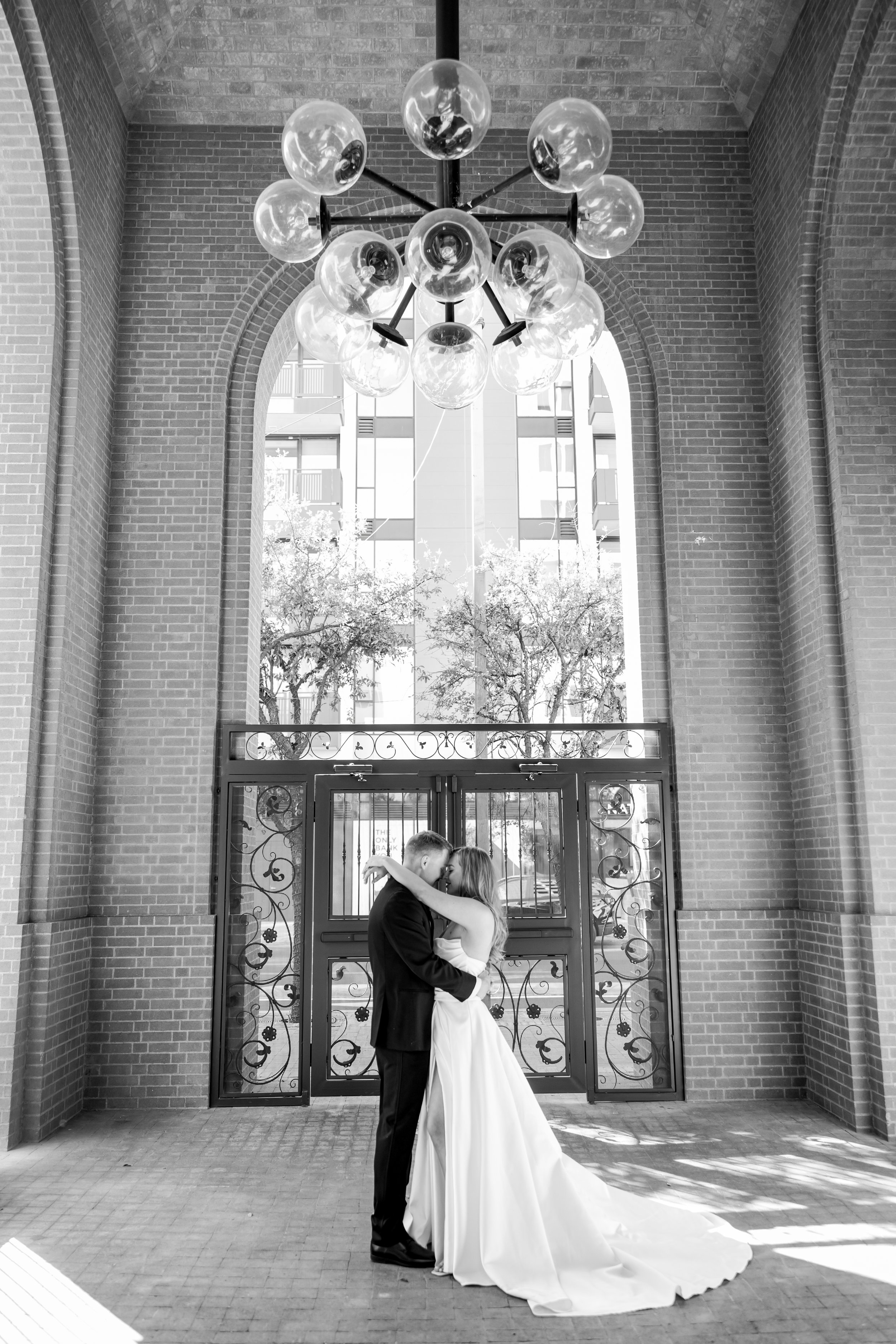 A black-and-white photo of a bride and groom embracing and kissing inside a brick building with large arched windows and a modern chandelier overhead.