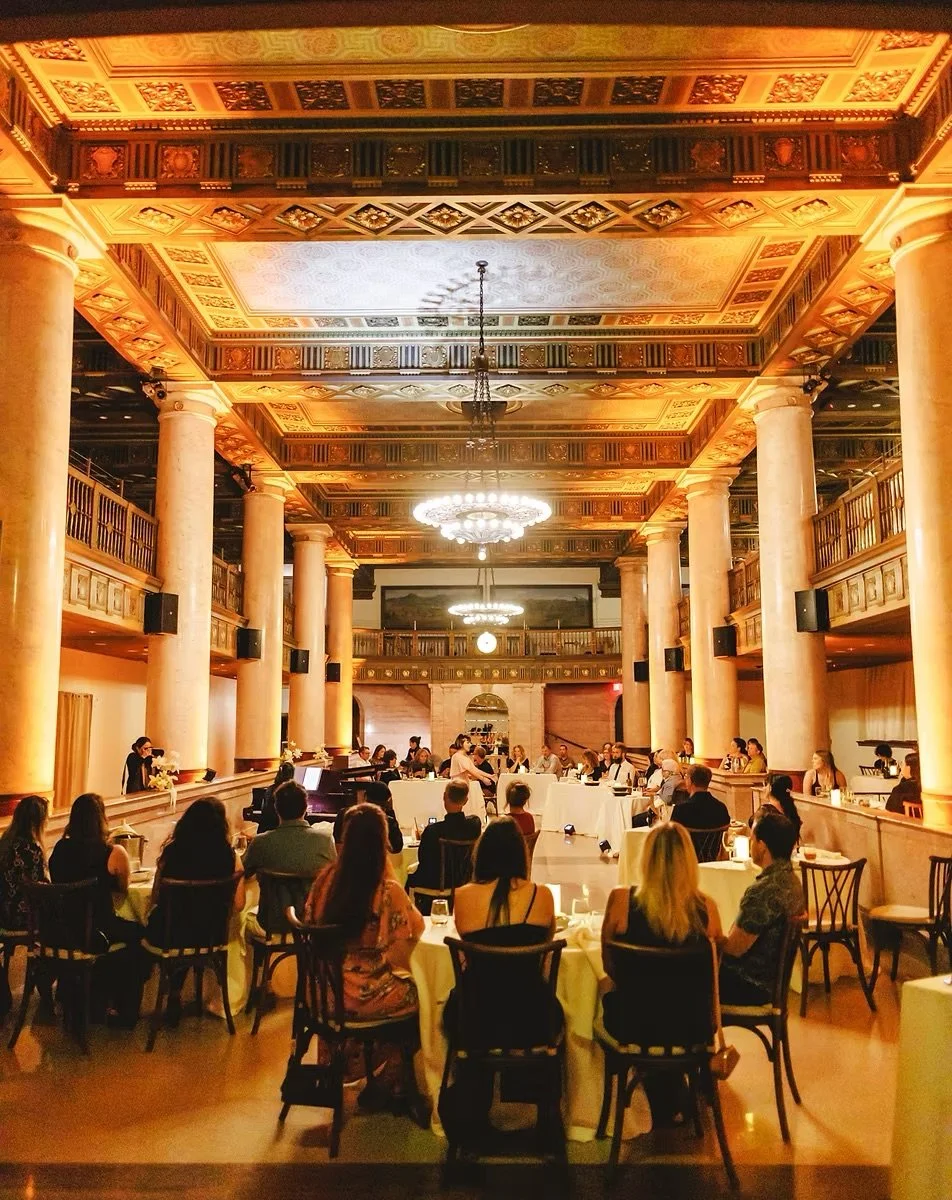 People seated around tables in an elegant ballroom with ornate ceiling, marble columns, and chandeliers during a performance or event.