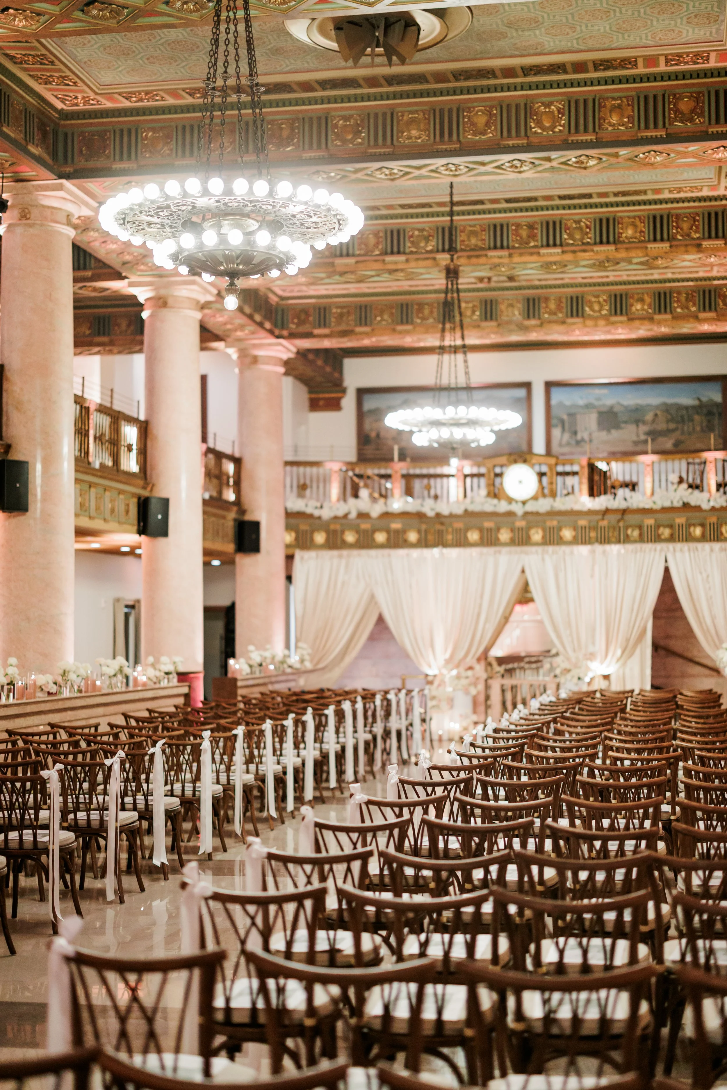 Elegant indoor wedding ceremony setup with rows of wooden chairs decorated with white ribbons, backdrop with cream curtains, ornate ceiling lighting, and decorative columns.