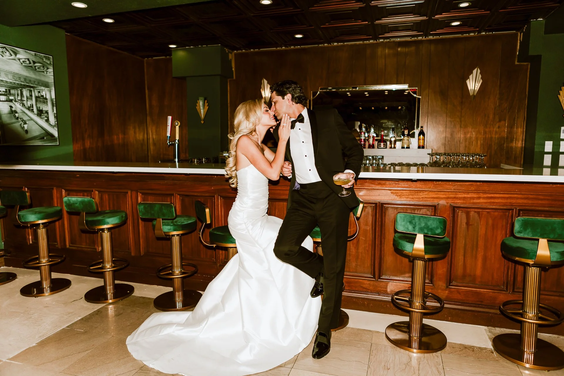 Bride and groom dancing at a bar, with the bride in a white wedding gown and the groom in a tuxedo, holding a drink, sharing a kiss.
