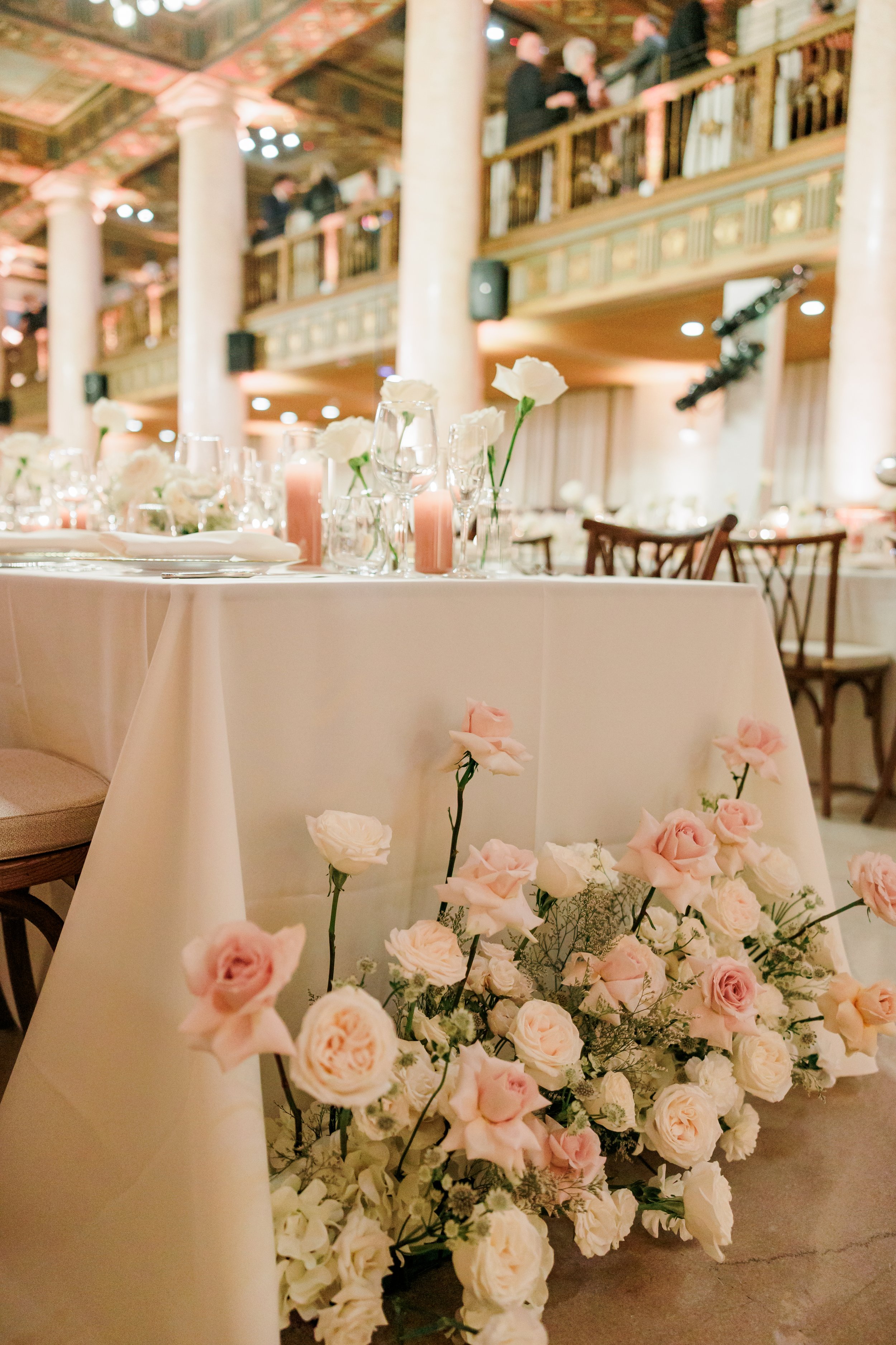 Elegant event hall decorated with pink and white roses, glassware, and candles on a table, with guests mingling on a balcony above.