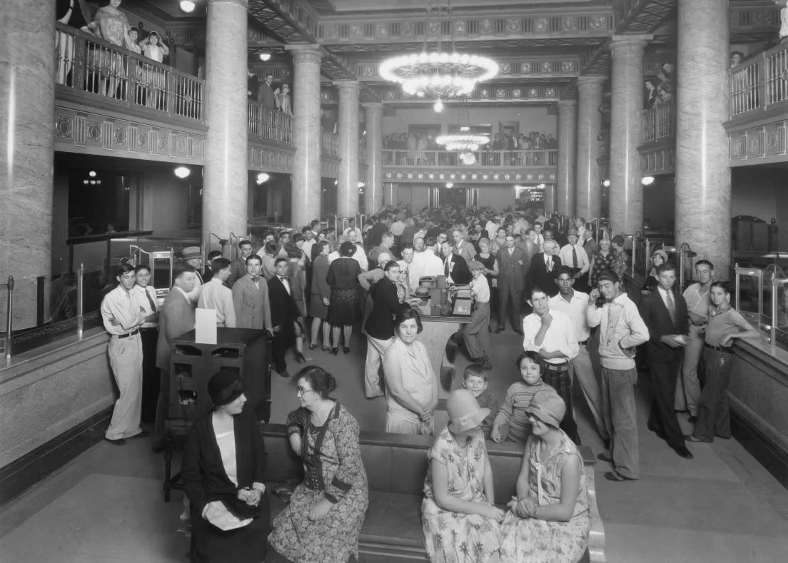 A busy theater lobby filled with people, including men, women, and children, during what appears to be the early 20th century. The scene shows a grand hall with tall columns, chandeliers, and balconies, with people standing in line and chatting.