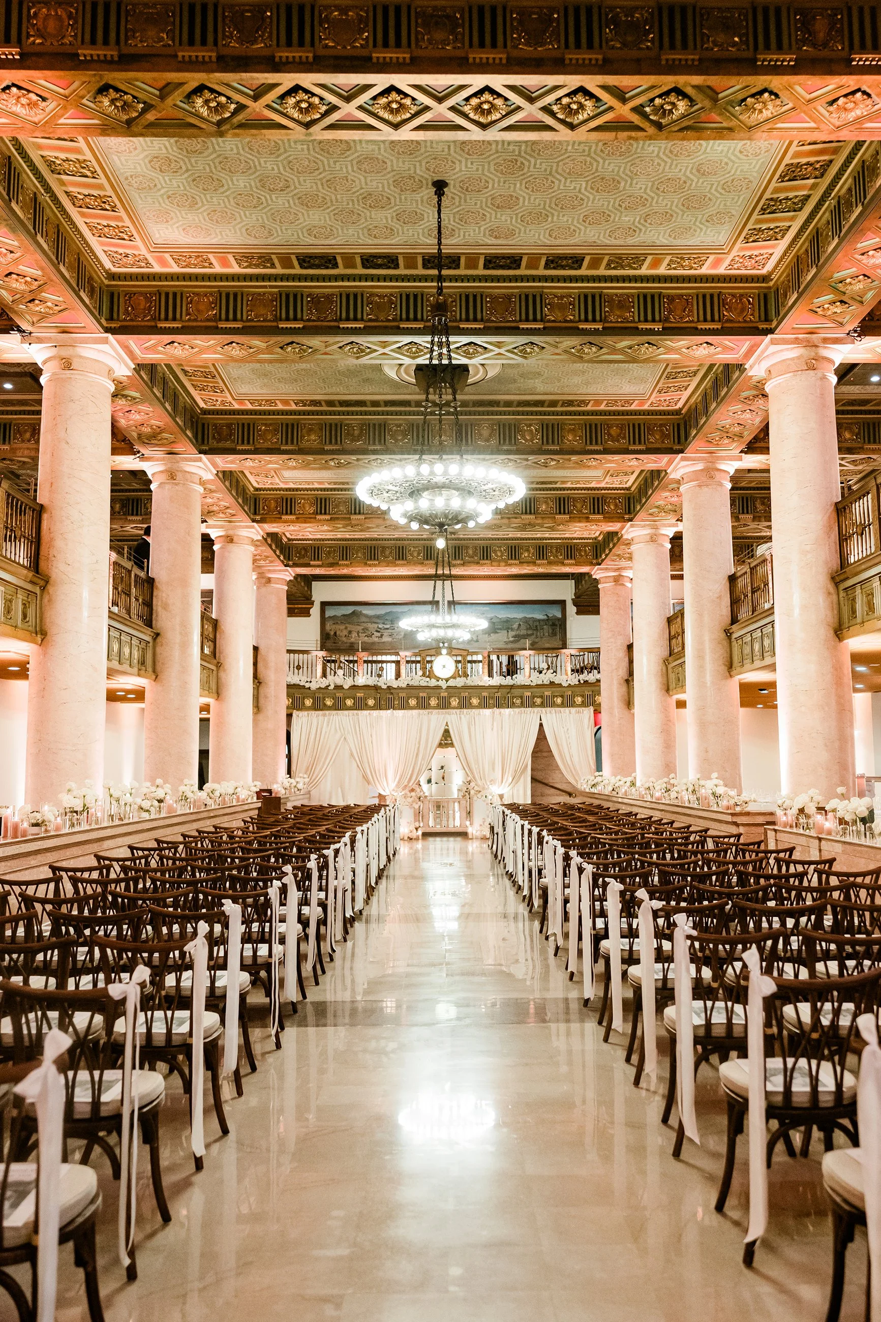 Elegant indoor wedding venue with rows of chairs, pink columns, ornate ceiling, and chandeliers, decorated with white flowers and drapery.