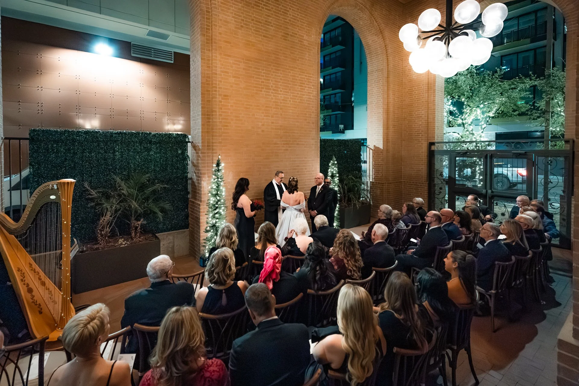 A wedding ceremony indoors with guests seated around the couple and officiant at the altar, decorated with small Christmas trees and plants, illuminated by a modern chandelier at night.