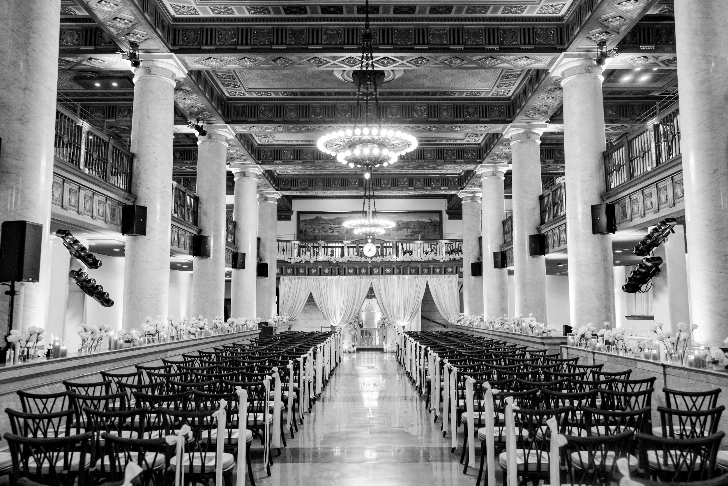 Black and white photo of an elegant event hall with high ceilings, large pillars, and chandeliers. Rows of chairs are set up on either side of a central aisle leading to a stage with curtains, flowers, and candles.