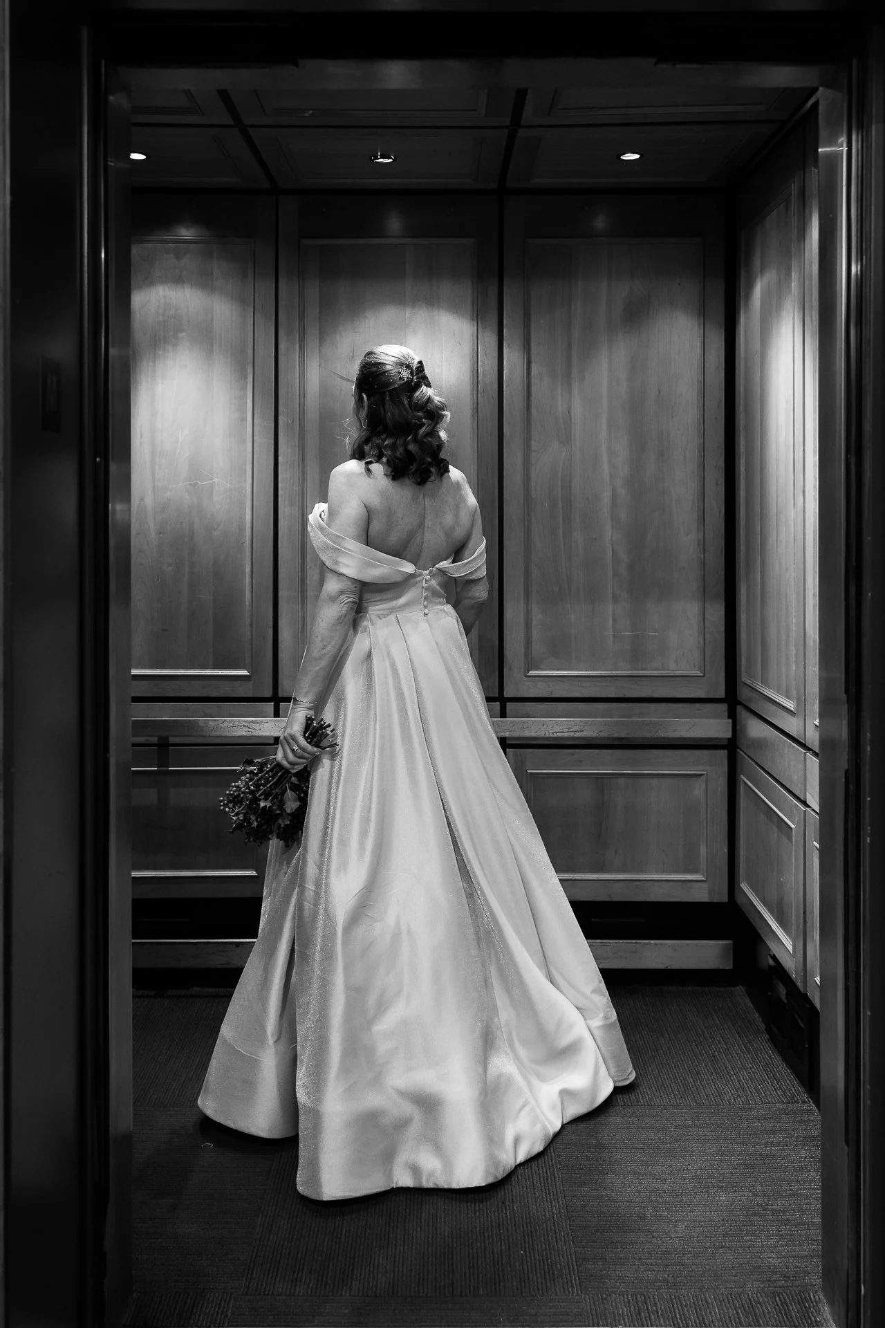 A woman in a wedding dress holding a bouquet in an elevator, seen from the back.