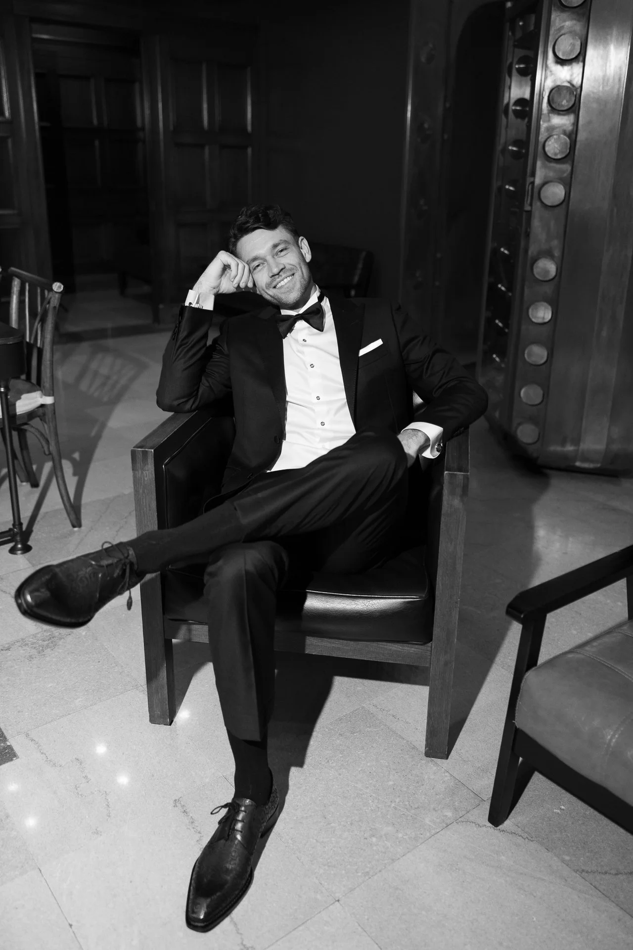 A man in a tuxedo sitting in an armchair, smiling with his leg crossed over another, in an elegant room.
