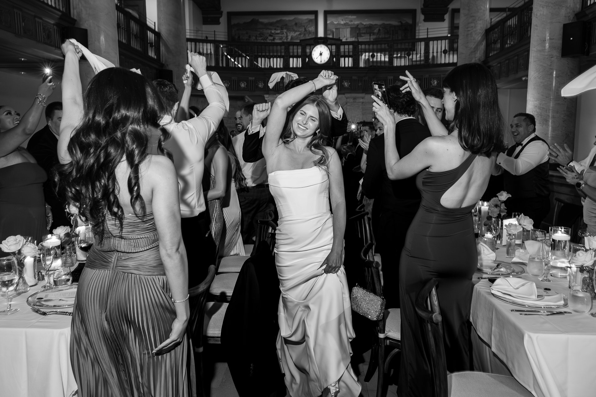 A woman in a strapless bridal gown dancing joyfully at a wedding reception, surrounded by guests who are celebrating and applauding.