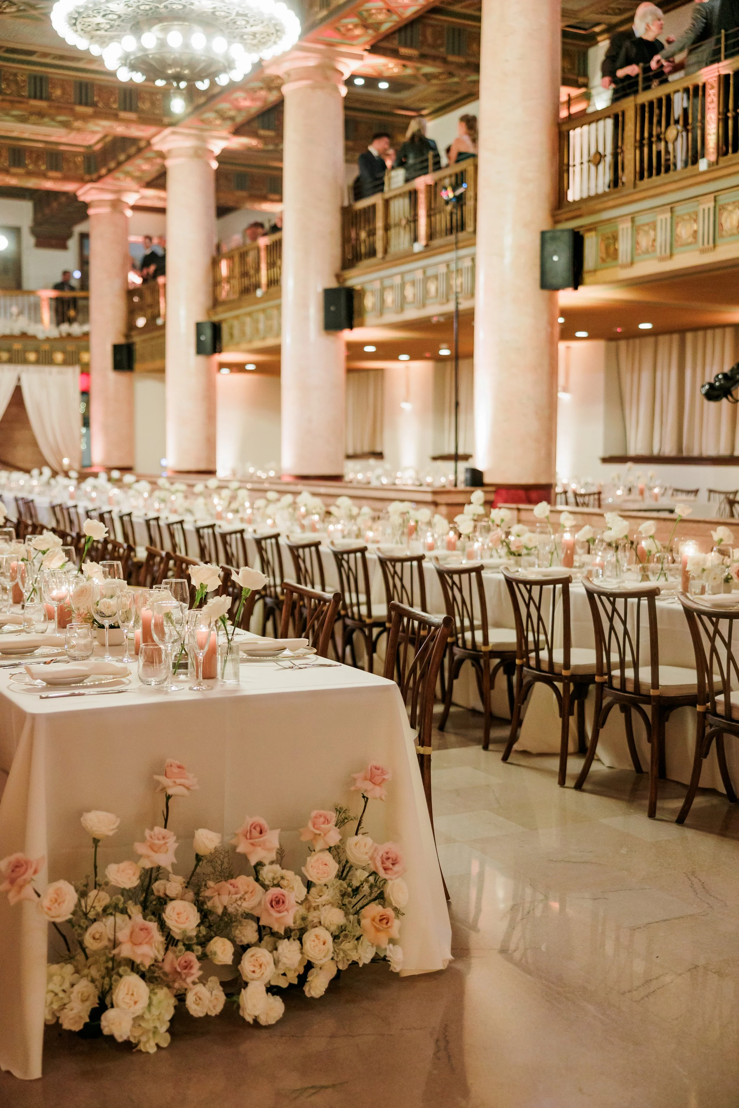 Elegant banquet hall decorated for a wedding reception with long tables, floral arrangements, and a chandelier, with people standing on a balcony overlooking the room.