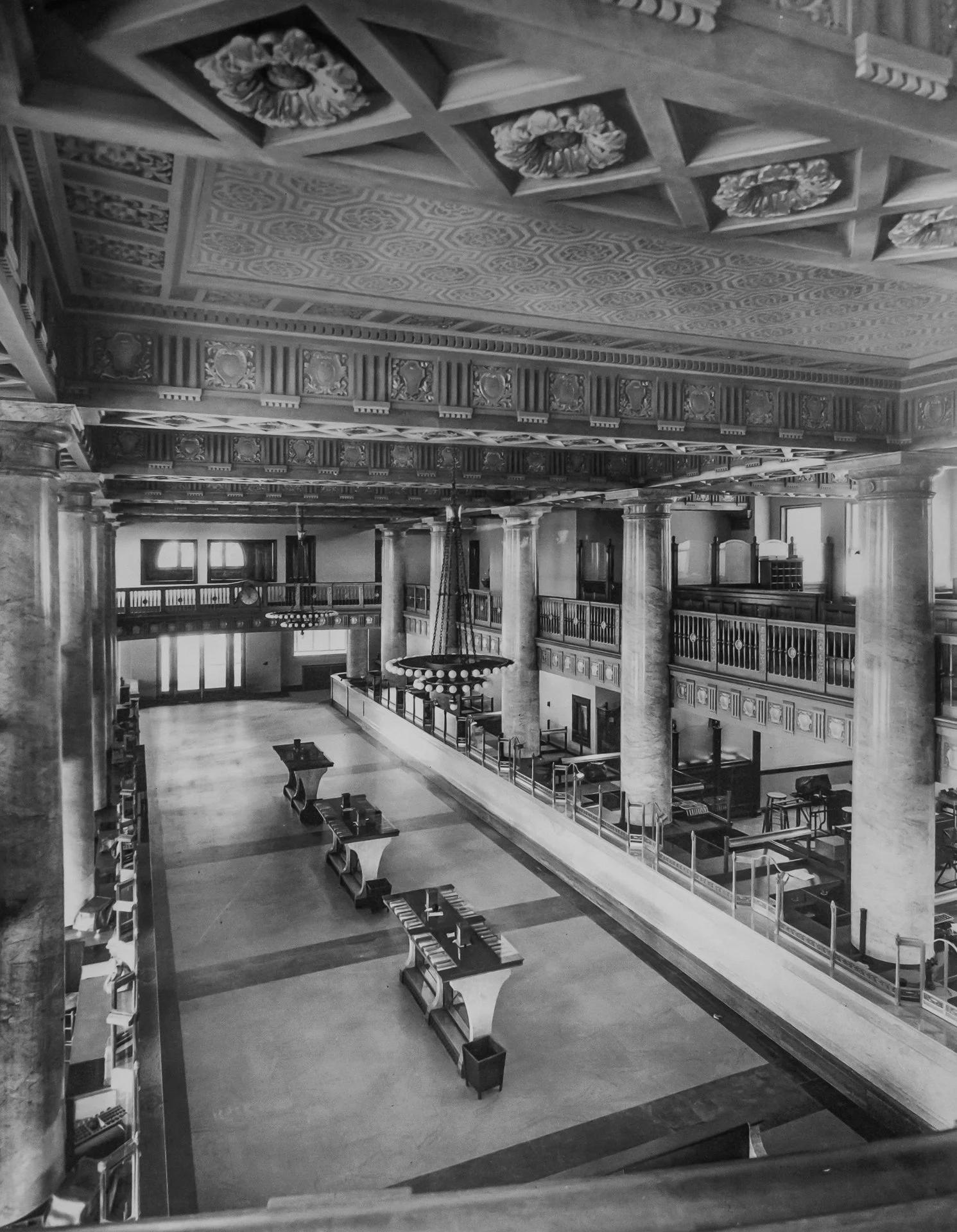 Black and white photo of an empty elegant grand hall with ornate ceiling, columns, and multiple levels of balcony. Several long tables and chairs are arranged along the hall. Large chandelier hangs from the ceiling.