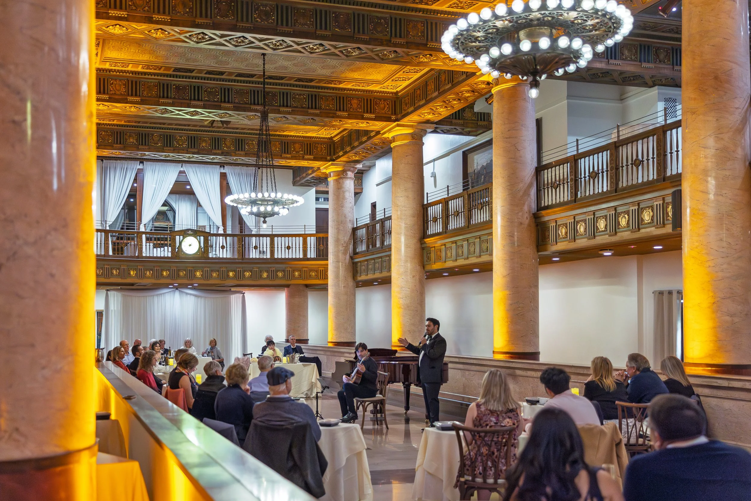 A man in a suit giving a speech while standing beside a seated musician playing a guitar at a formal event inside an ornate ballroom with high ceilings, large columns, chandeliers, and hanging curtains, with an audience seated at round tables.