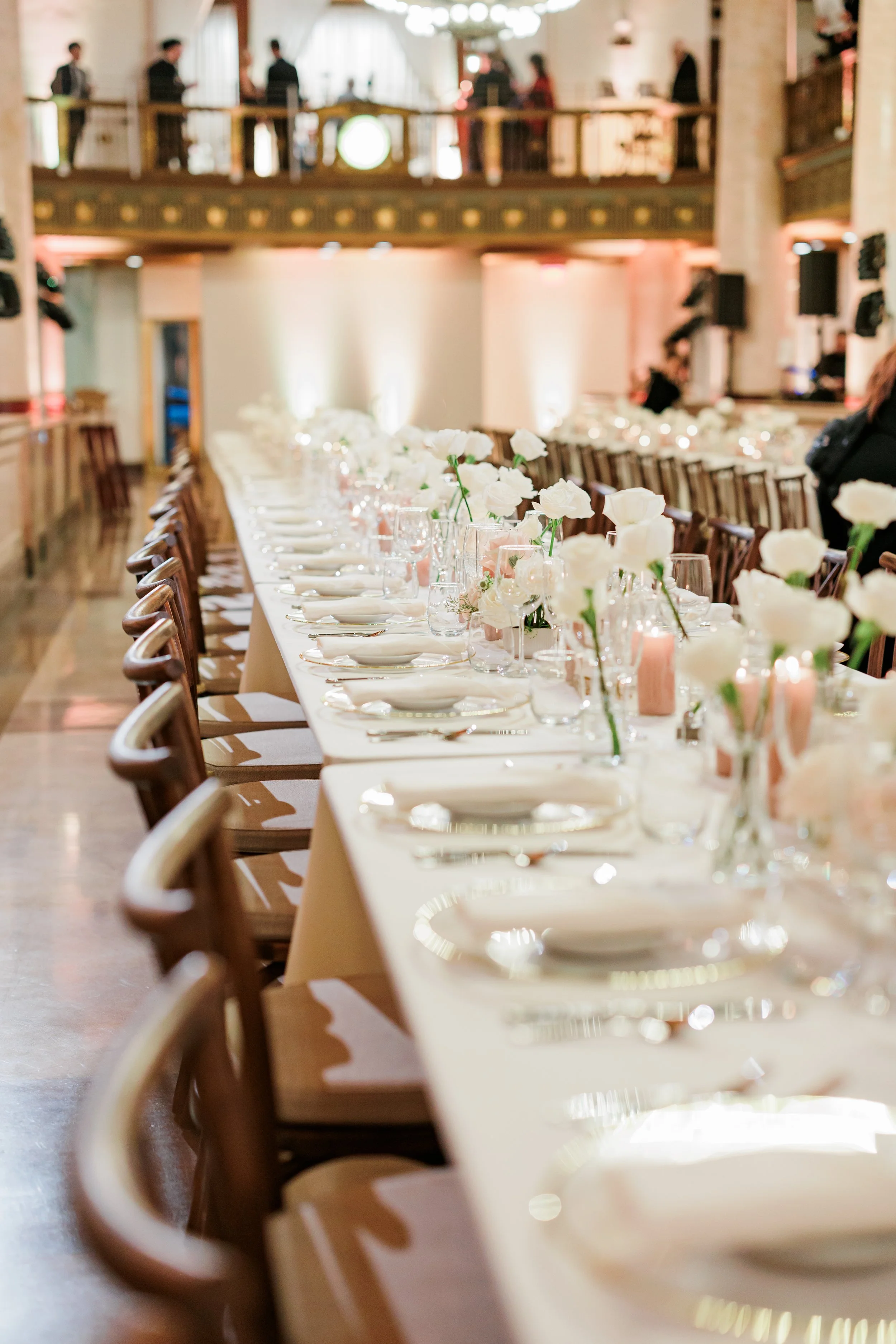 Elegant banquet table set for a formal event with white flowers, candles, and glassware, in a decorated hall with balcony.