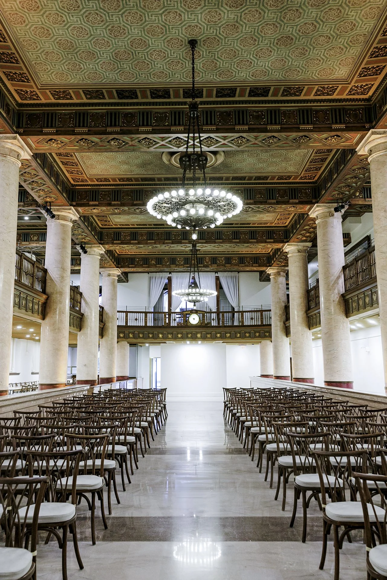 An elegant, spacious hall with rows of chairs for an event. Features tall marble columns, ornate ceiling with intricate patterns, and large chandeliers hanging from the ceiling. A balcony with a clock overlooking the hall is visible in the background.