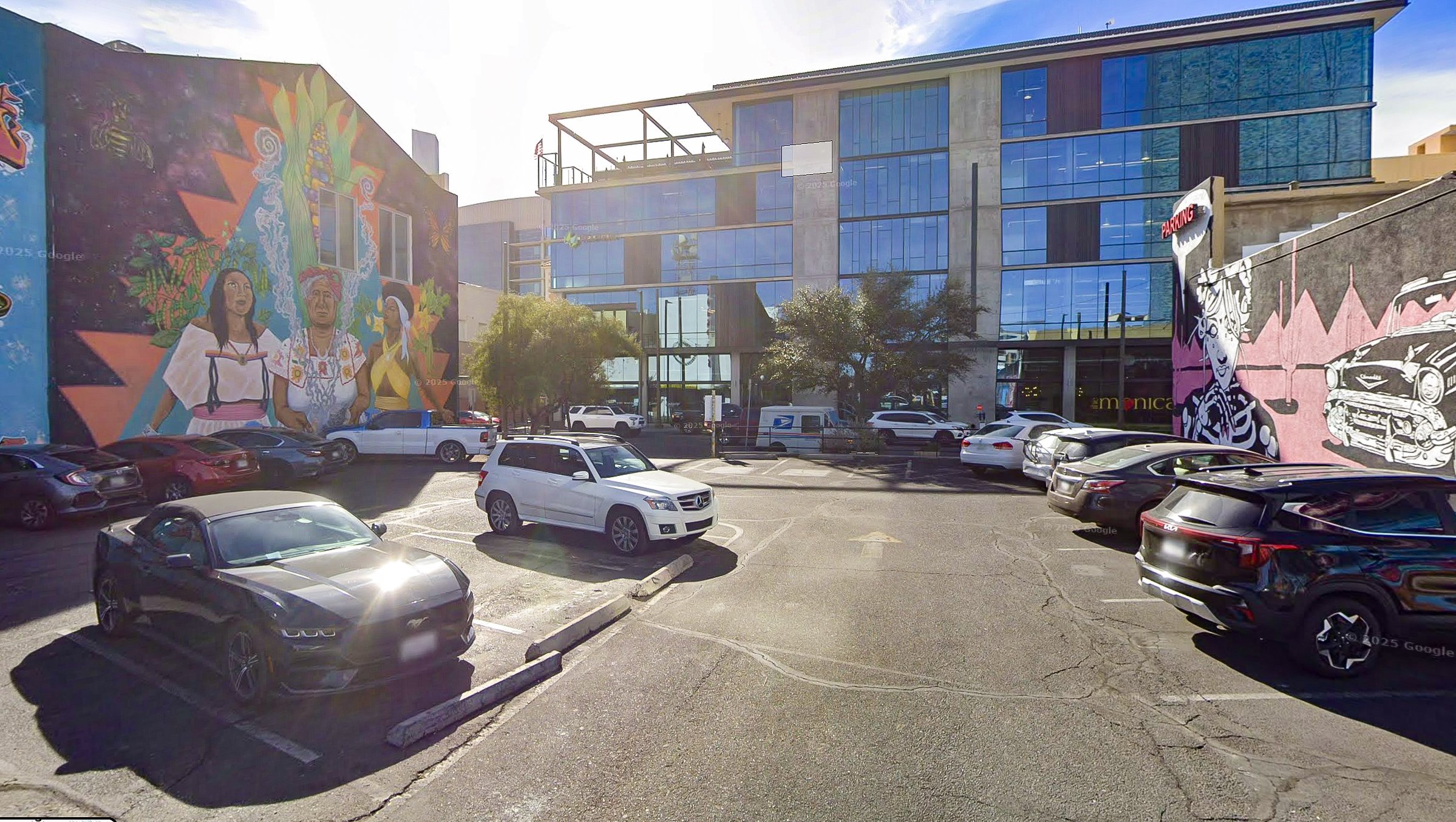A parking lot in front of a modern glass building with colorful murals on the walls depicting people, vehicles, and artistic designs.
