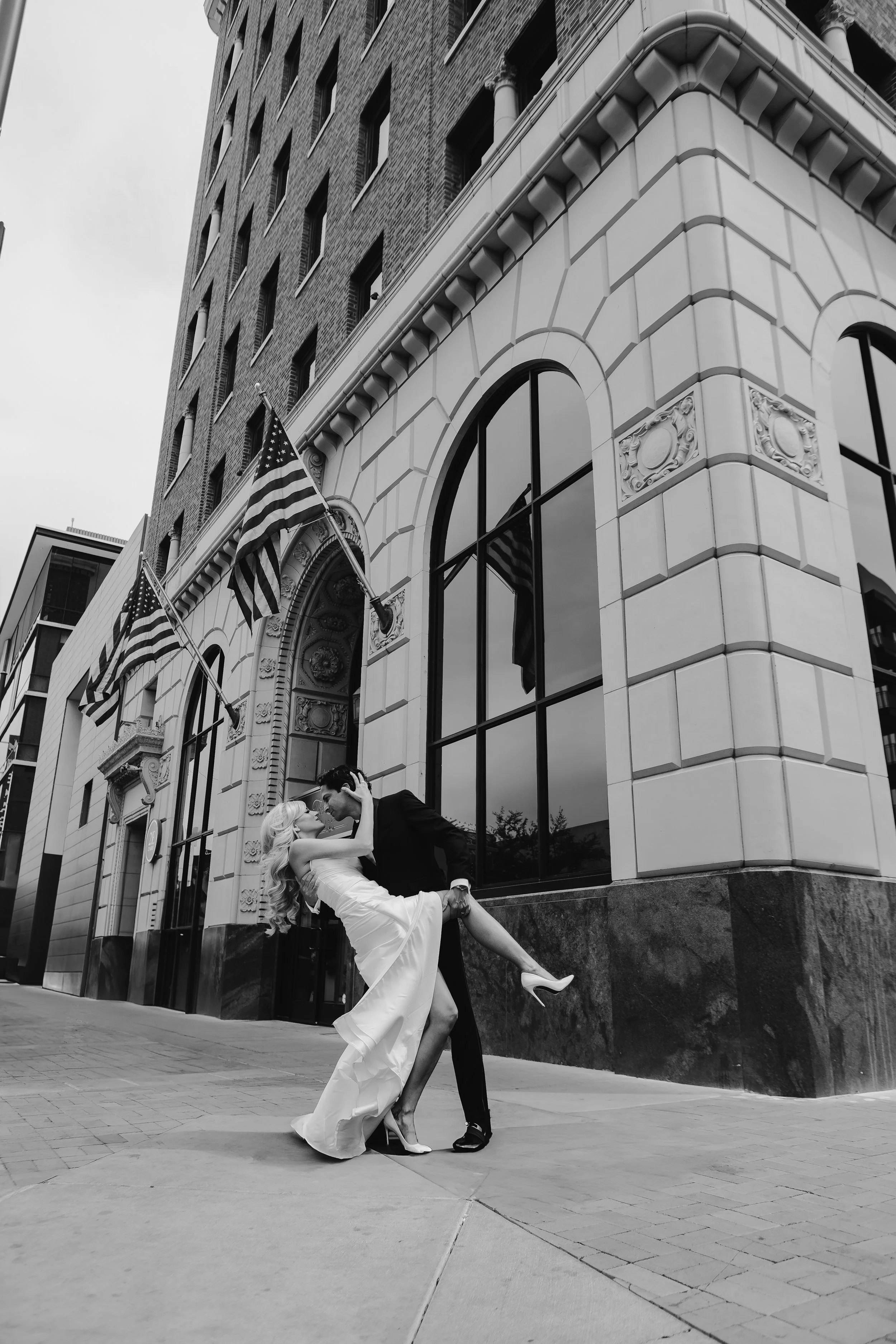 Black and white photo of a couple dancing on a city sidewalk in front of a large ornate building with American flags. The woman in a white dress is being dipped by the man in a dark suit, with her leg raised and heels visible.