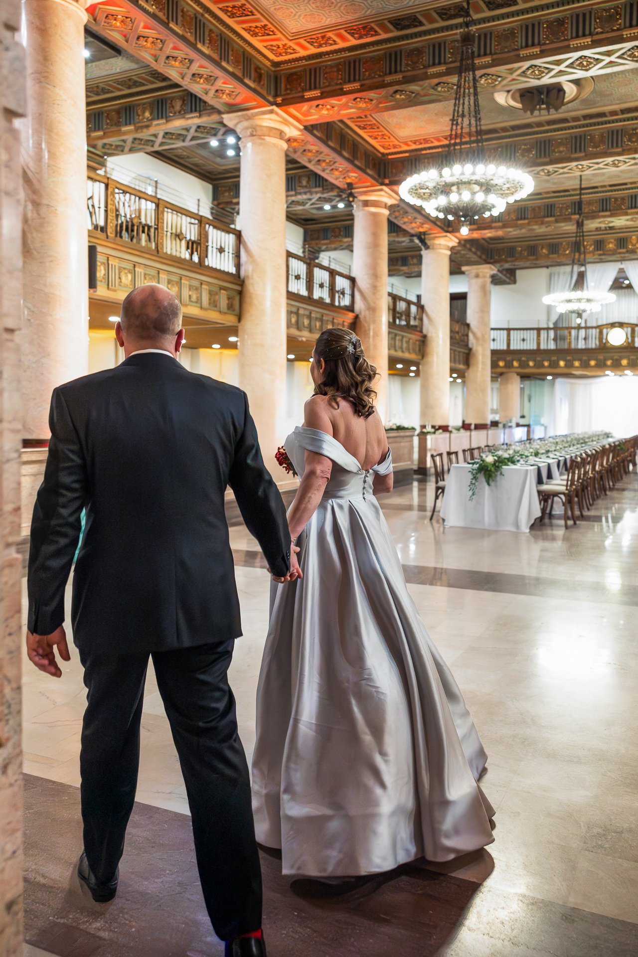 A bride and groom holding hands as they walk into a decorated wedding hall with high ceiling, large columns, and elegant chandeliers.