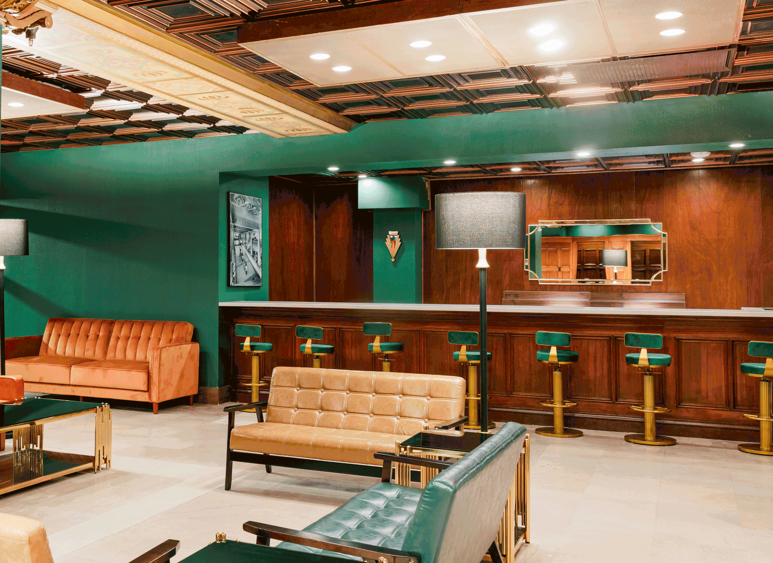A stylish hotel bar with green walls, wooden accents, and retro furniture including a tan leather sofa, an orange velvet sofa, and green bar stools with brass bases. A large mirror, artwork, and modern lamps add to the decor.