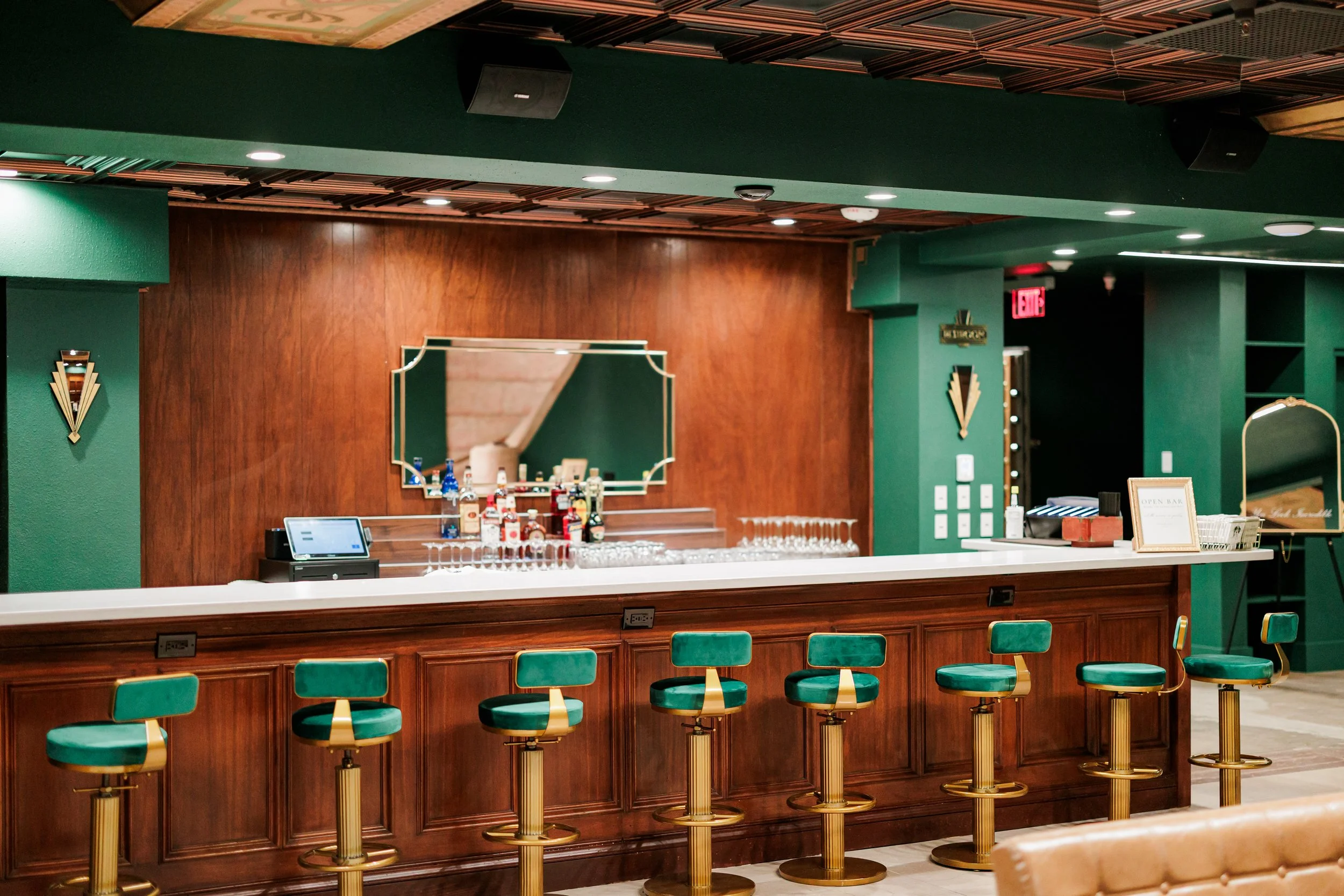An elegant bar with a polished wooden counter, green velvet bar stools with gold bases, and a mirror behind the bar reflected in the background. The bar is stocked with bottles of liquor, barware, and a touchscreen point-of-sale system. The decor fea
