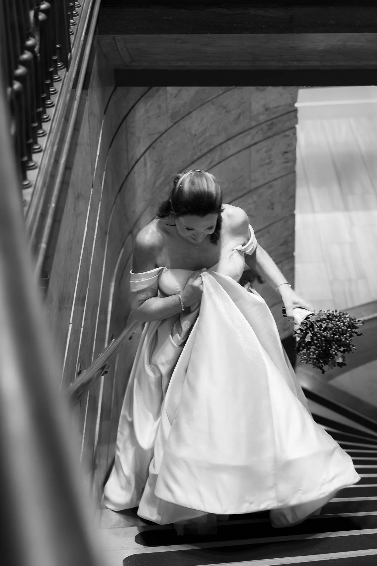 A bride dressed in a wedding gown descending a staircase holding a bouquet of flowers, viewed from above in black and white.