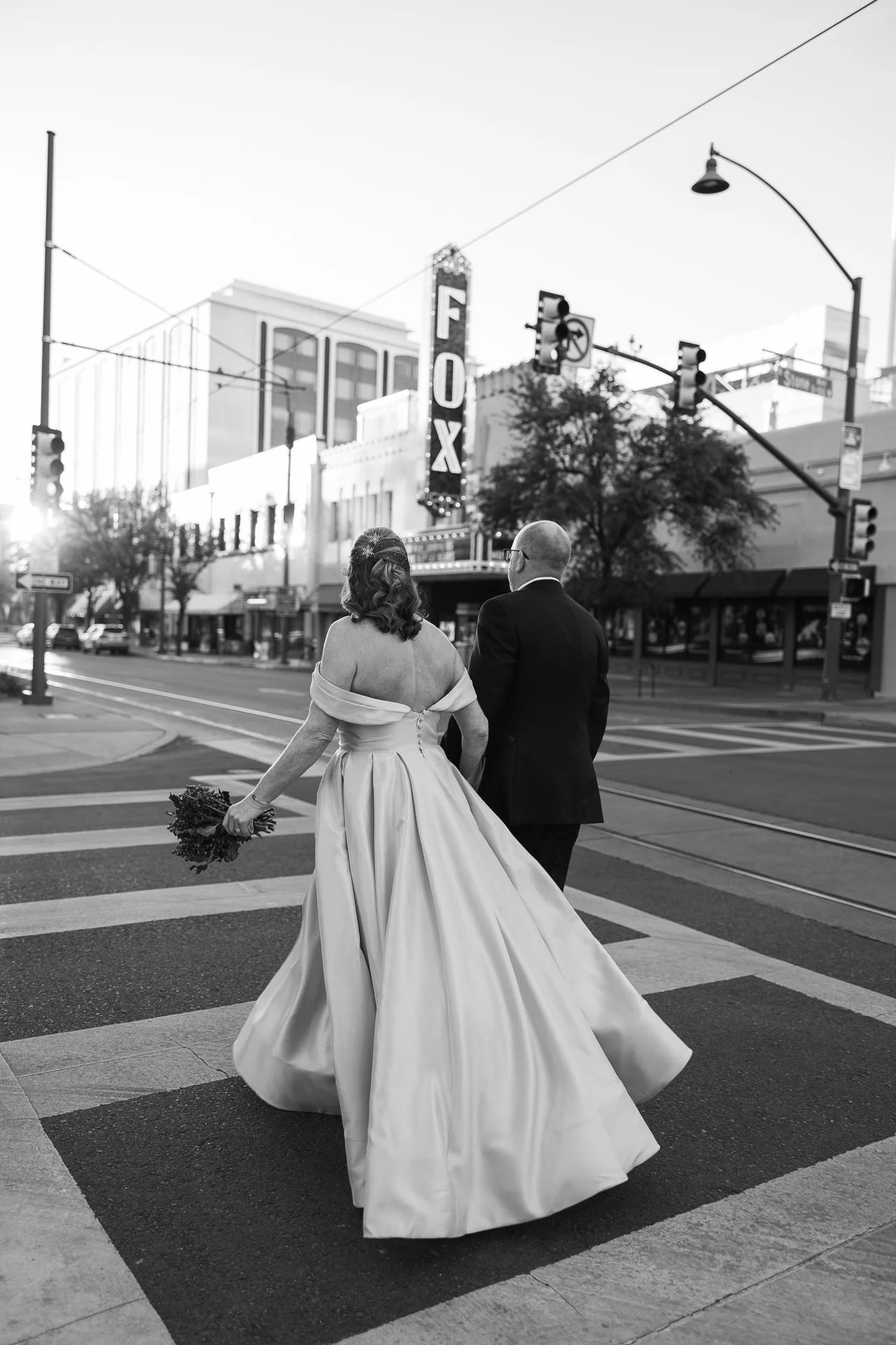 A bride in a wedding gown holding a bouquet and a man in a tuxedo crossing a city street on a crosswalk in black and white.