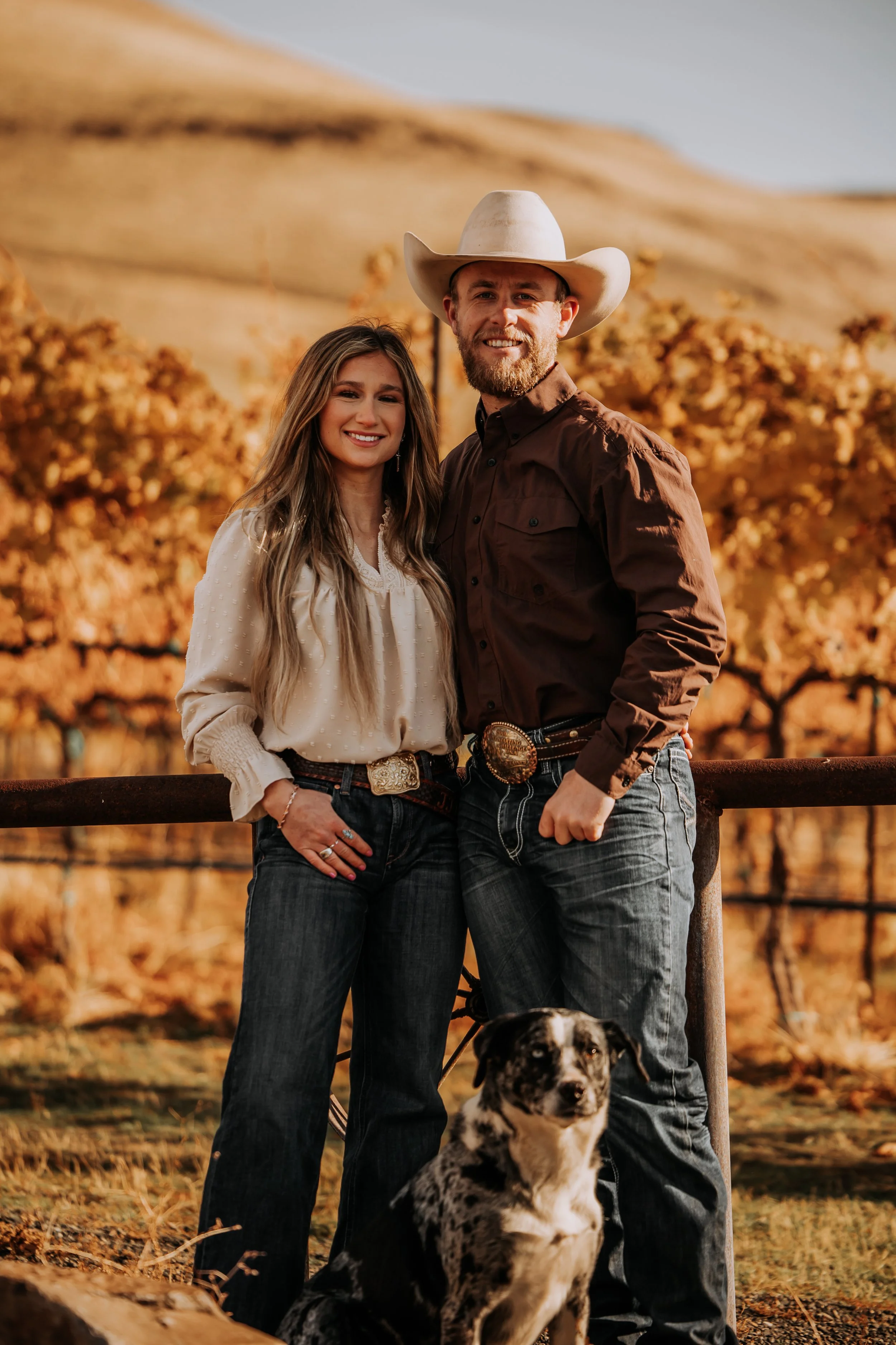 A couple dressed in cowboy attire standing outdoors with a dog, autumn trees, and rolling hills in the background.