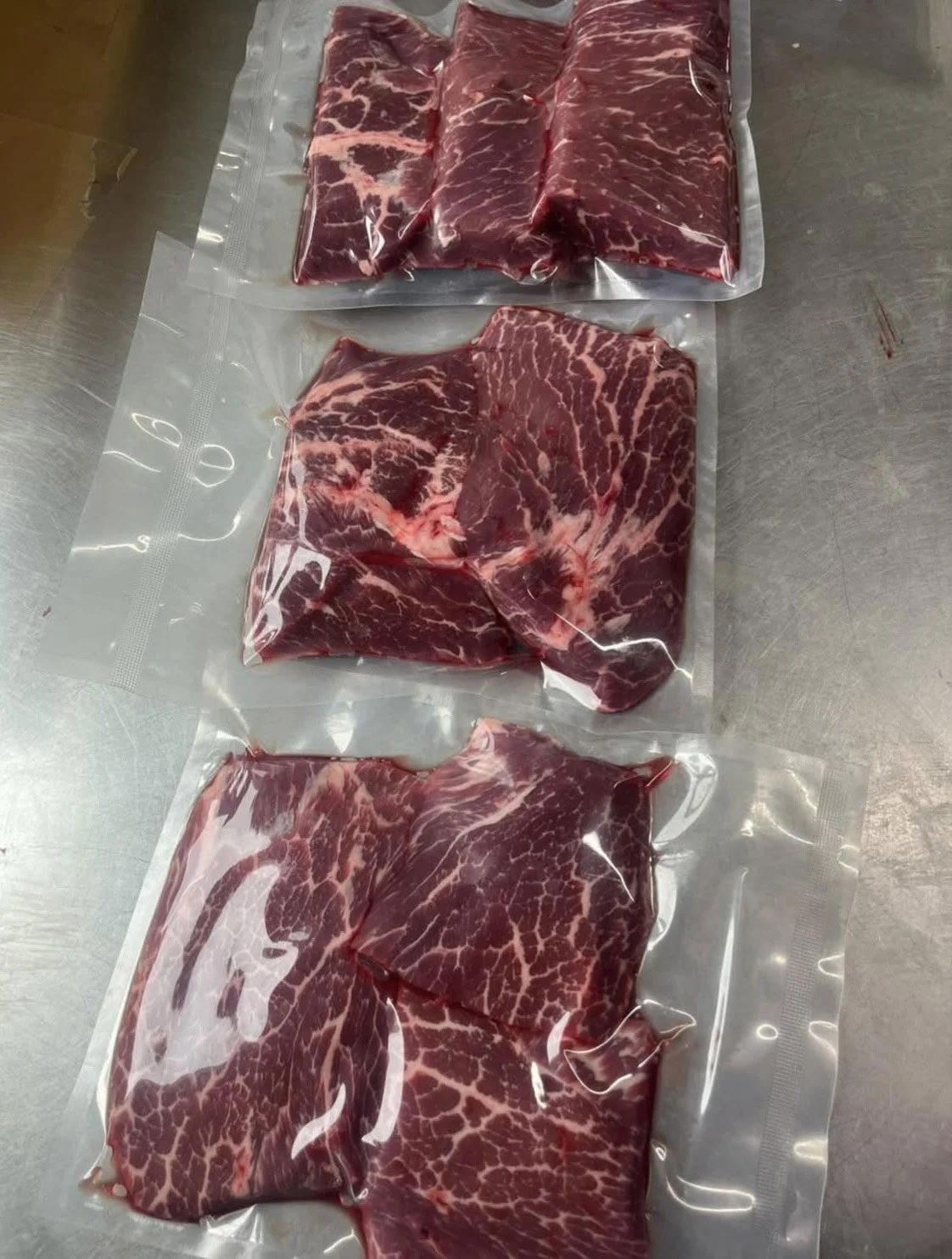 Three vacuum-sealed packages of raw beef with marbling, placed on a stainless steel surface.