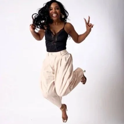 Joyful woman with long black hair jumping in the air, wearing a black top and beige pants, smiling and making peace signs with her hands.