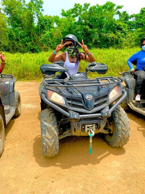Woman in sunglasses and face mask making peace signs while sitting on an all-terrain vehicle on a dirt path, with green foliage in the background.