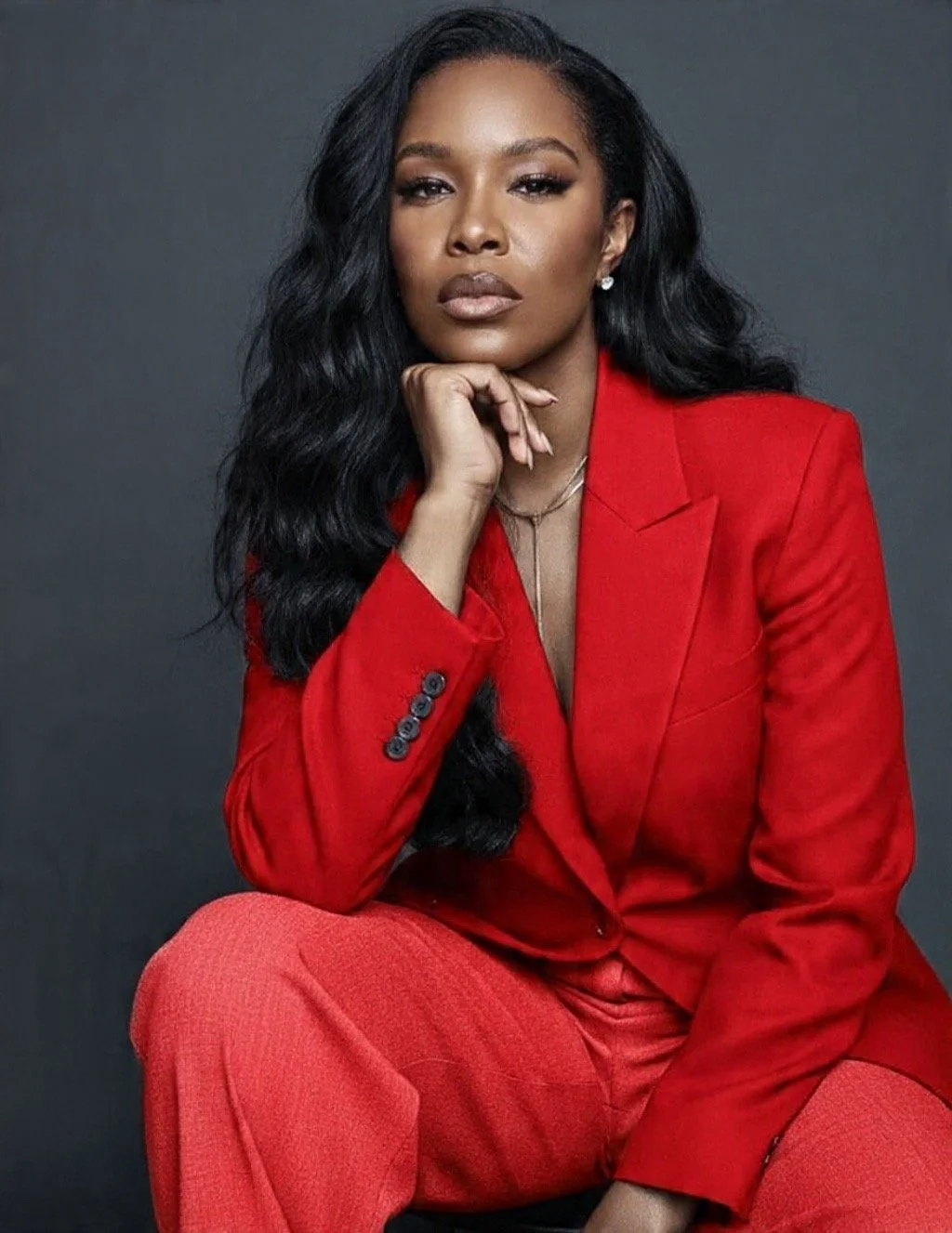 A woman with long wavy black hair wearing a red blazer and red pants, sitting against a gray background with her chin resting on her hand.