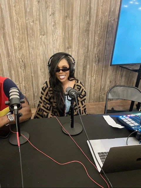 A woman with long dark hair wearing sunglasses and headphones, smiling, sitting at a table with a microphone and audio equipment, in a room with wood-paneled walls.