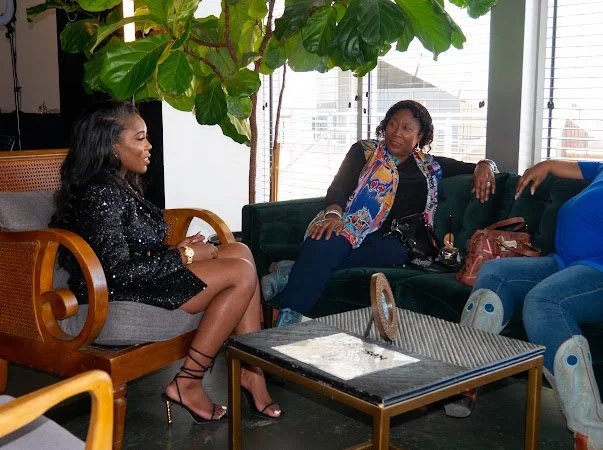 Three women sitting and talking in a stylish indoor lounge with a large green plant and tall windows in the background.