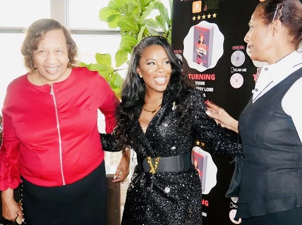 Three women engaged in conversation at an indoor event, with a promotional display in the background.