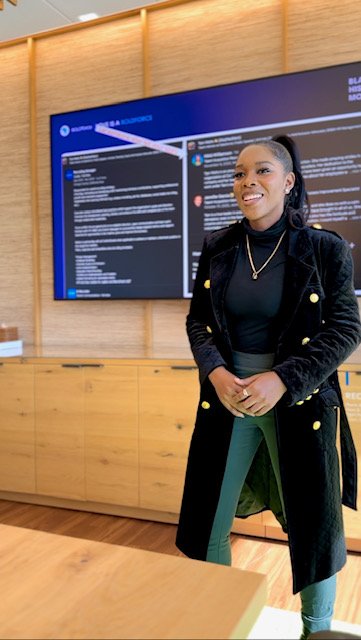 A woman standing and smiling in front of a digital screen displaying information in an indoor setting.