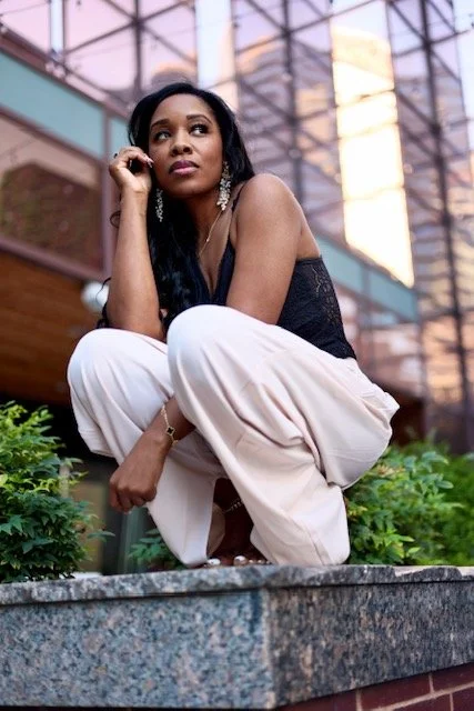 A woman with long black hair, dressed in a black top and light-colored pants, crouching on a stone surface outdoors with greenery, glass buildings, and a sunset sky in the background.