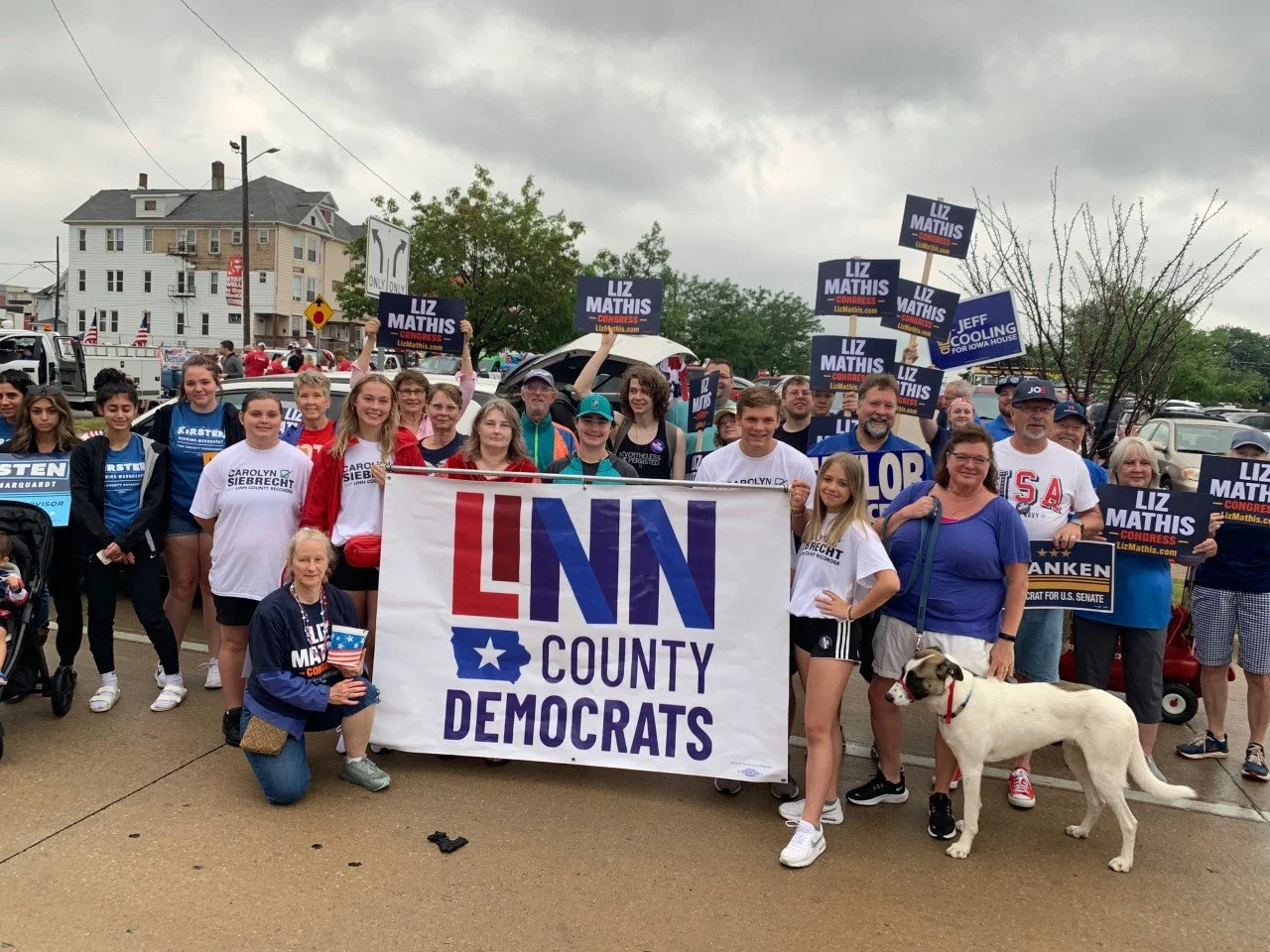 Group of people holding a large sign that reads "LNN County Democrats," with some holding campaign signs for Liz Mathis and others. There is a woman kneeling in front holding a small American flag and a white dog with black spots is standing beside a woman in a blue shirt. The background shows a cloudy sky and residential buildings.