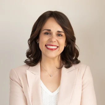 A woman with shoulder-length brown hair smiling in a beige blazer and white top against a plain background.