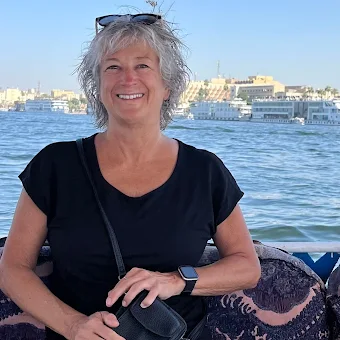 A smiling woman with gray hair in a black shirt, sitting by a body of water, with city buildings in the background.