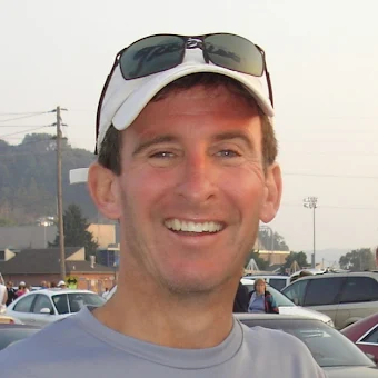 A man smiling outdoors, wearing a white cap with sunglasses on top, in a parking lot with cars and power lines in the background.