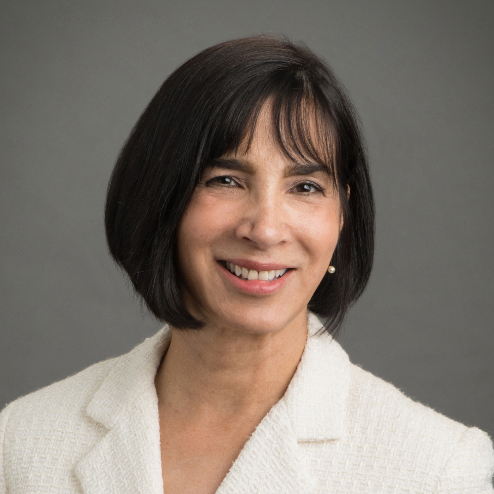 A woman with short dark hair, smiling, wearing a cream blazer and pearl earring against a gray background.