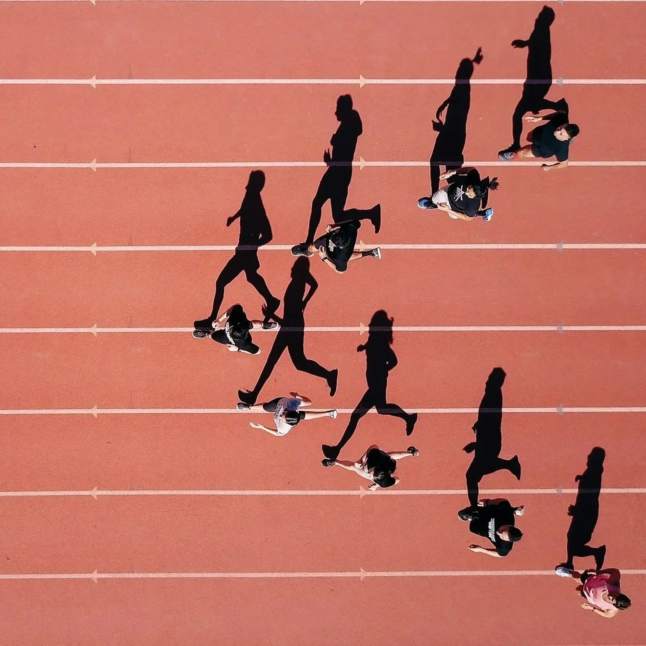 Shadows of multiple people running on an outdoor track, captured from above during daylight.